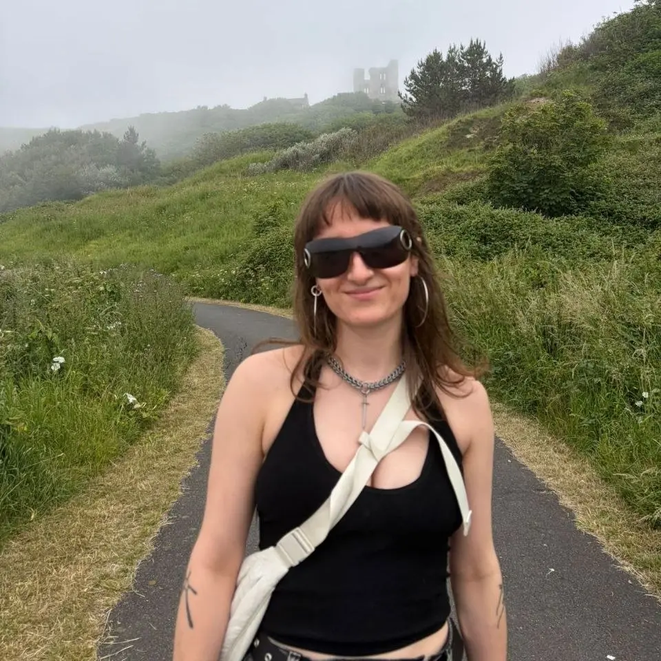 Seren Oakley stands on a road in front of hills. The fog is dense behind her and she's wearing sunglasses and smiling.